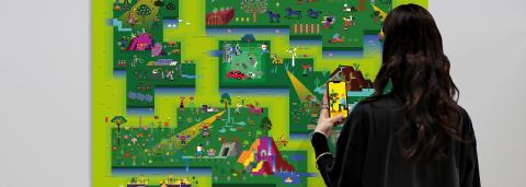 Visuel de BlockSmog, on y voit une personne qui joue sur son téléphone au jeu intéractif qui compose l'exposition, elle se trouve devant une des fresques