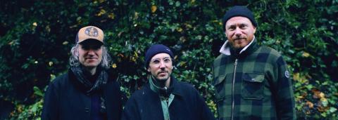 photo de trois hommes (membres du groupe), debout devant de la verdure (arbres et terre)