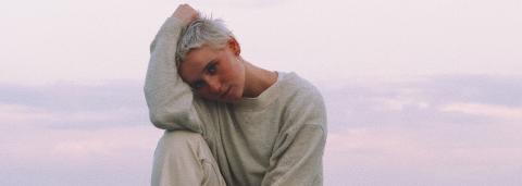 Kat White, les cheveux court, blond platine, posant en pull amble écru, assise sur un rocher, au bord de la mer, la lumière rosé du matin en fond.