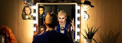 ne personne est assise de dos devant un miroir entouré d’ampoules allumées, dans une loge. Son reflet montre un visage transformé avec un maquillage drag très expressif, yeux soulignés de noir et lèvres accentuées. Elle porte une perruque beige sur la tête, en préparation d’une performance. Le miroir est décoré de découpages de portraits de stars (dont Elvis Presley et d'autres icônes hollywoodiennes)