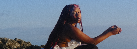 Odoneila en photo au bord de la mer, les cheveux longs tressés, avec des mêches rouges  portant une robe blanche et un pantalon noir