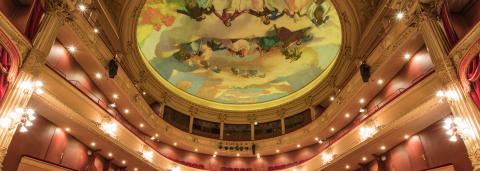 Intérieur d’un opéra à l’architecture classique, vu depuis la scène : plusieurs balcons ornés de dorures et de velours rouge entourent la salle, illuminés par de nombreux luminaires. Au plafond, une vaste fresque circulaire peinte représente des figures et des nuages dans des tons pastel