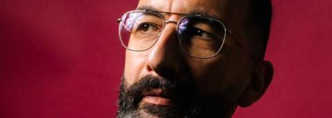 Portrait de Marwan Mohammed, le fond derrière lui est rouge uni, on le voit de profil portant des lunettes et un pull noir
