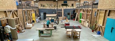 Photographie qui montre l'intérieur des ateliers de la coopérative Comme un établi. L'atelier est tout en bas et il y a une mezzanine sur le côté supérieur de la photo. On y voit différents plans de travail, avec deux personnes travaillant au centre. Sur la gauche, une personne est en train de monter une porte en bois sur une structure.