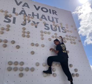 Une personne montre le mur braille, en haut du mur est écrit la phrase "Va voir là-haut si j'y suis"