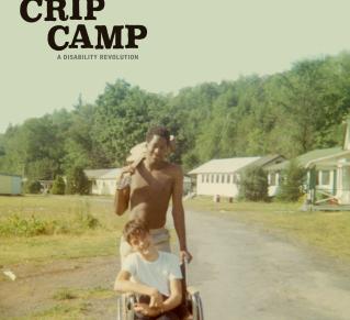 Affiche du film "CRIP CAMP" Deux jeunes garçons sont au centre de l’image, sur une route d’un camp de vacances entouré de bâtiments blancs et de nature verdoyante. L’un est assis dans un fauteuil roulant, souriant, vêtu d’un t-shirt blanc et d’un pantalon noir. L’autre, debout derrière lui, torse nu et tenant une serviette sur l’épaule, pousse le fauteuil.