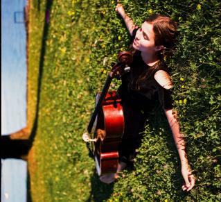 Photo de Dom La Nena dans l'herbe verte, allongée avec son violoncelle