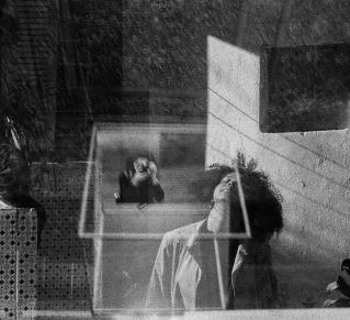 Superposition de deux photo, on y voit au premier plan une femme les cheveux crépus, les yeux fermés, levé un peu la tête, derrière elle, une personne apparait en reflet la prenant en photo. Sur le côté droit, on apperçoit un bâtiment.