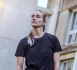 Portrait de la danseuse Annaelle Toussaere, les cheveux blond cendrés, attachées, portant un t shirt noir et un jeans de couleur brute les bras tombant