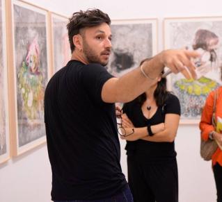 Photo portrait de Romuald Jandolo portant un t shirt noir des lunettes dans la main gauche devant une exposition