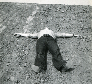 photographie en noir et blanc d'une personne allongée sur le dos sur un terrainen pente, rocheux, les bras en croix, son visage est cachée par l'angle de prise de vue, le ciel occupe la partie dupérieur de l'image.