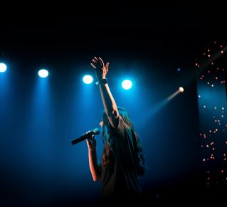 Une personne au cheveux long chante, un micro à la main et lève l'autre main, derrière on voit des lumière de scène bleu