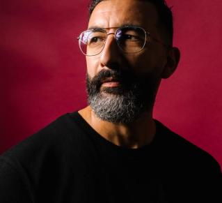 Portrait de Marwan Mohammed, le fond derrière lui est rouge uni, on le voit de profil portant des lunettes et un pull noir.