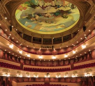 Intérieur d’un opéra à l’architecture classique, vu depuis la scène : plusieurs balcons ornés de dorures et de velours rouge entourent la salle, illuminés par de nombreux luminaires. Au plafond, une vaste fresque circulaire peinte représente des figures et des nuages dans des tons pastel