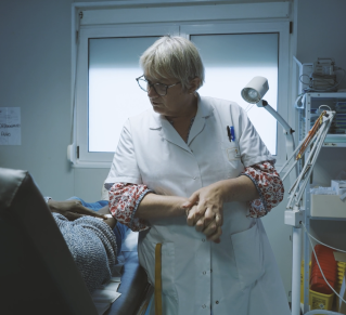 Visuel du film "Carnets de soignants : qu'est-ce que bien soigner ?" on y voit une soignante en blouse blnache portant des lunettes de vue dans une salle blanche parler avec un patient dont on devine uniquement les bras.