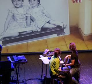 Photo issue du spectacle où on apperçoit deux comédiennes, brunes, asisses dos au public. Elles regardent la projection sur grand écran une photo de famille, sur laquelle deux petites filles sourient.