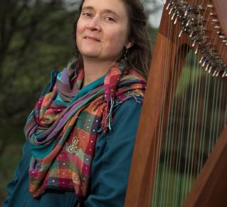 Portrait de Hoëla Barbedette, debout habillée avec une veste bleu et une écharpe de couleur à côté de sa harpe