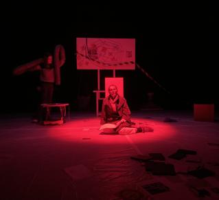 Photo prise lors d'une résidence de création, on y voit deux comédiennes sur scène, la llumière est sombre rouge. Un des comédiennes est au centre de l'image, elle lit un texte. L'autre comédienne se trouve derrière au fond de la scène