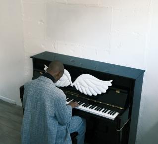 Un personne, vue de dos, est assise devant un piano droit noir et en joue. Une paire d'ailes blanches est posée sur le cylindre du piano
