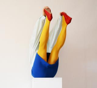 Photographie en couleur, sur un fond gris, d'une personne assise sur un socle blanc, les jambes levées vers le haut, portant une chemise blanche, un short bleu, des collants jaunes opaques et des escarpins rouges.