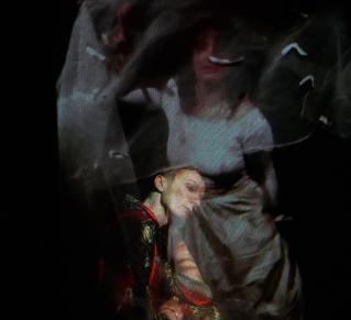 Visuel de "Rouge, amère fantasmagorie", on y voit deux figures humaines, créant un effet de flou et fantomatique. La personne à l'arrière-plan porte une robe blanche avec un visage partiellement visible, créant une impression de mouvement. Au premier plan, une autre figure est habillée de vêtements aux couleurs vives, principalement rouge et doré. La figure en arrière plan penche la tête sur le coté, donnant une impression de fragilité ou de réflexion.