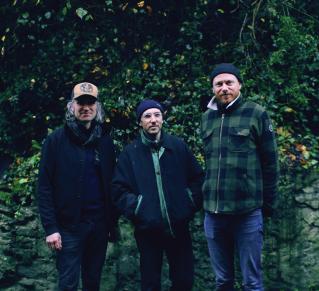 photo de trois hommes (membres du groupe), debout devant de la verdure (arbres et terre)