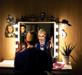 Une personne est assise de dos devant un miroir entouré d’ampoules allumées, dans une loge. Son reflet montre un visage transformé avec un maquillage drag très expressif, yeux soulignés de noir et lèvres accentuées. Elle porte une perruque beige sur la tête, en préparation d’une performance. Le miroir est décoré de découpages de portraits de stars (dont Elvis Presley et d'autres icônes hollywoodiennes)
