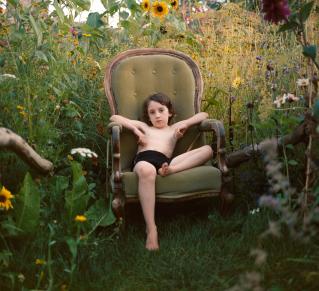 Visuel The Garden, un enfant sur un fautueil vert, fixe la caméra est entouré de verdure, haute herbes et fleurs