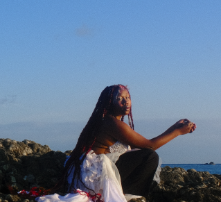 Odoneila en photo au bord de la mer, les cheveux longs tressés, avec des mêches rouges  portant une robe blanche et un pantalon noir
