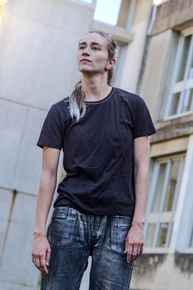 Portrait de la danseuse Annaelle Toussaere, les cheveux blond cendrés, attachées, portant un t shirt noir et un jeans de couleur brute les bras tombant