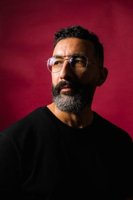 ortrait de Marwan Mohammed, le fond derrière lui est rouge uni, on le voit de profil portant des lunettes et un pull noir.