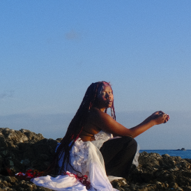 Odoneila en photo au bord de la mer, les cheveux longs tressés, avec des mêches rouges  portant une robe blanche et un pantalon noir