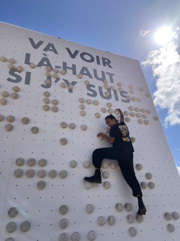 Une personne montre le mur braille, en haut du mur est écrit la phrase "Va voir là-haut si j'y suis"