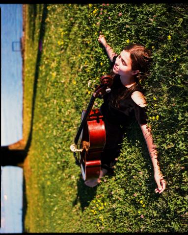 Photo de Dom La Nena dans l'herbe verte, allongée avec son violoncelle