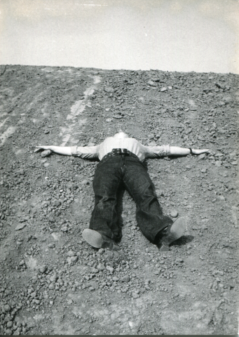 photographie en noir et blanc d'une personne allongée sur le dos sur un terrainen pente, rocheux, les bras en croix, son visage est cachée par l'angle de prise de vue, le ciel occupe la partie dupérieur de l'image.