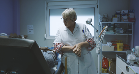 Visuel du film "Carnets de soignants : qu'est-ce que bien soigner ?" on y voit une soignante en blouse blnache portant des lunettes de vue dans une salle blanche parler avec un patient dont on devine uniquement les bras.