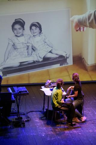 Photo issue du spectacle où on apperçoit deux comédiennes, brunes, asisses dos au public. Elles regardent la projection sur grand écran une photo de famille, sur laquelle deux petites filles sourient.