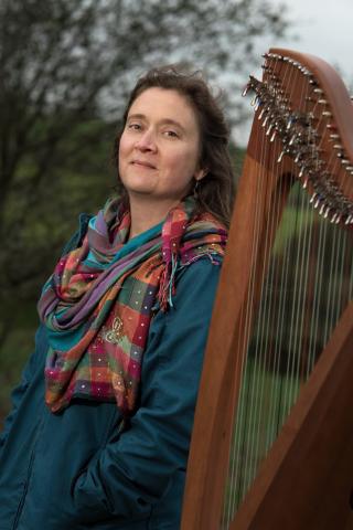Portrait de Hoëla Barbedette, debout habillée avec une veste bleu et une écharpe de couleur à côté de sa harpe