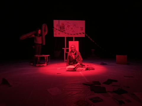 Photo prise lors d'une résidence de création, on y voit deux comédiennes sur scène, la llumière est sombre rouge. Un des comédiennes est au centre de l'image, elle lit un texte. L'autre comédienne se trouve derrière au fond de la scène