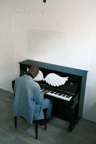 Un personne, vue de dos, est assise devant un piano droit noir et en joue. Une paire d'ailes blanches est posée sur le cylindre du piano