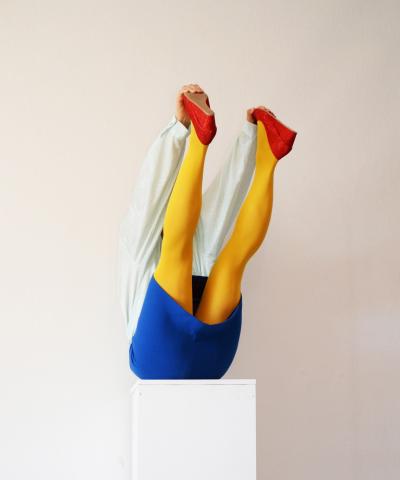 Photographie en couleur, sur un fond gris, d'une personne assise sur un socle blanc, les jambes levées vers le haut, portant une chemise blanche, un short bleu, des collants jaunes opaques et des escarpins rouges.