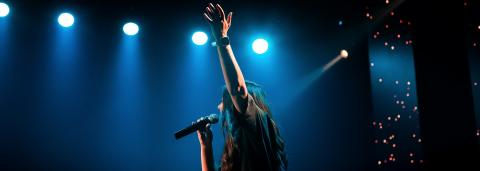 Une personne au cheveux long chante, un micro à la main et lève l'autre main, derrière on voit des lumière de scène bleu