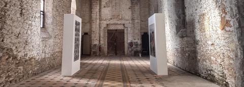 C'est une photo de l'intérieur de l'espace d'exposition de Noir Brillant. Les murs sont en pierre avec une grande hauteur sous plafond. Au fond, on aperçoit une porte en bois. Le sol est en carrelage à motif noir et blanc. Au centre, on aperçoit deux blocs blanc qui se font fasse avec des photos.