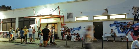 Il s'agit d'une photo extérieure du bâtiment du Jardin Moderne. On y aperçoit la devanture du bâtiment. Un groupe d'une dizaine de personnes sont juste devant l'entrée, sous un porche rouge. On aperçoit également une personne sur un banc sur la droite avec un tag sur le mur.