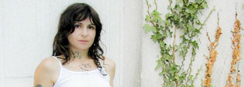 Portrait de Louïse Papier, les cheveux noirs, mi long, avec une frange, portant un t-shirt blanc et des tatouages