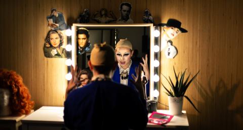Une personne est assise de dos devant un miroir entouré d’ampoules allumées, dans une loge. Son reflet montre un visage transformé avec un maquillage drag très expressif, yeux soulignés de noir et lèvres accentuées. Elle porte une perruque beige sur la tête, en préparation d’une performance. Le miroir est décoré de découpages de portraits de stars (dont Elvis Presley et d'autres icônes hollywoodiennes