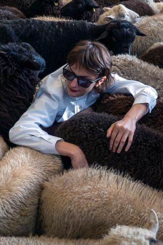 Visuel faire troupeau, photo de Marion Thomas, (au centre)  portant des lunettes noires de soleil, entourée de d'un troupeau de moutons 