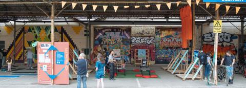 Photo des Halles en communs, on y voit un espace sous un préau, décoré avec une girlande en papier. L'espace est tagué, avec des graffitis, on y voit une exposition et plusieurs personnes regardent cette exposition.