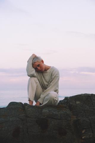 Kat White cheveux blond platine sur un rocher, au bord de la mer avec en arrière plan l'aurore, un ciel très claire, rosée