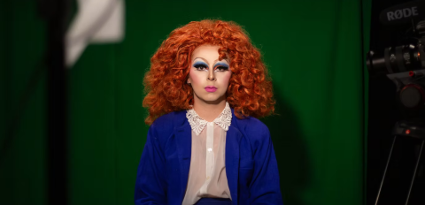 Portrait de Lucas Rahon en drag-queen, avec une pérruque rousse aux cheveux bouclés arrivant aux épaules, du fard bleu et orange sur les yeux, du rouge à lèvre rose, portant un costume bleu et une chemise blanche avec un col en dentelle